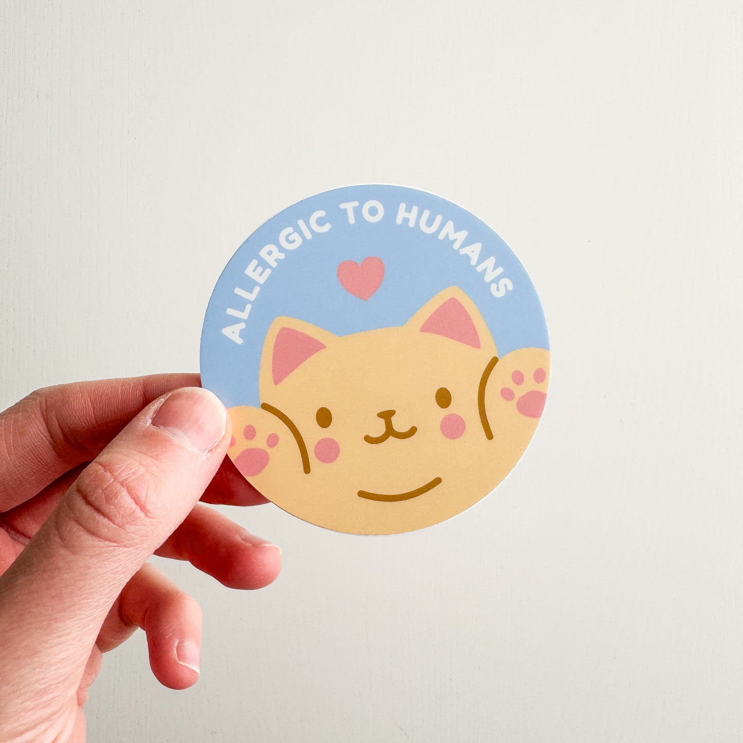 Circular sticker with a cat illustration and text 'Allergic to Humans' held by a hand on a light background