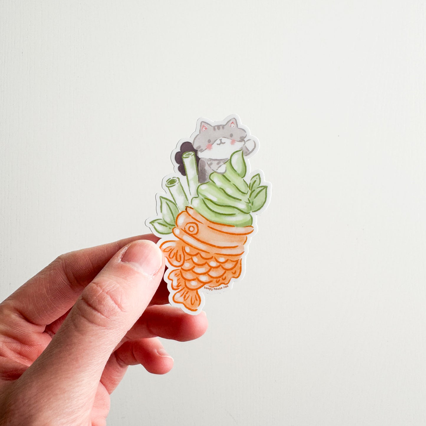 Hand holding a sticker of a Matcha Taiyaki with a cat inside on a plain background