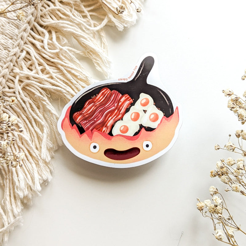 Calcifer sticker featuring a cute flame character in a skillet with bacon and eggs, matte waterproof finish.