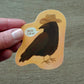 Hand holding a vinyl sticker of a crow wearing a cowboy hat saying "yee-caw" on an orange background.