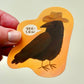 Hand holding a vinyl sticker of a black crow wearing a cowboy hat saying yee-caw on an orange background