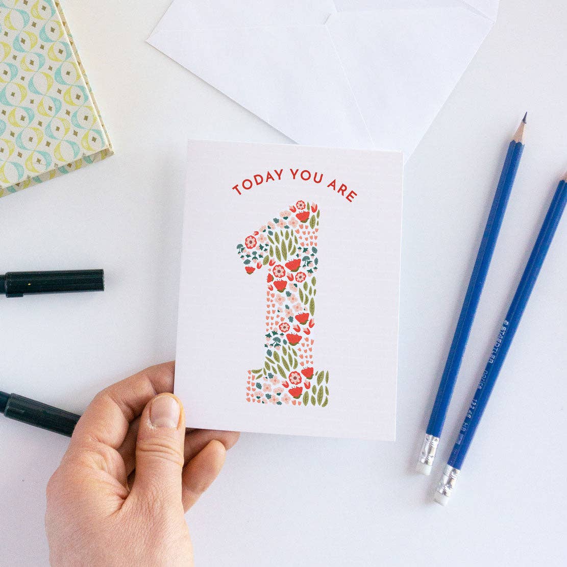 Hand holding a card with floral number '1' design and text 'Today you are 1' on a white surface with pens and an envelope.