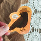Hand holding a vinyl sticker of a crow wearing a cowboy hat saying "yee-caw" on an orange background.
