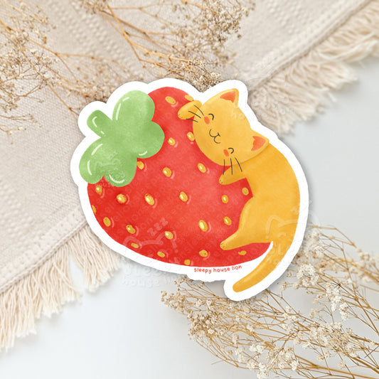 Sticker of a cat on top of a strawberry on a textured surface with dried flowers.