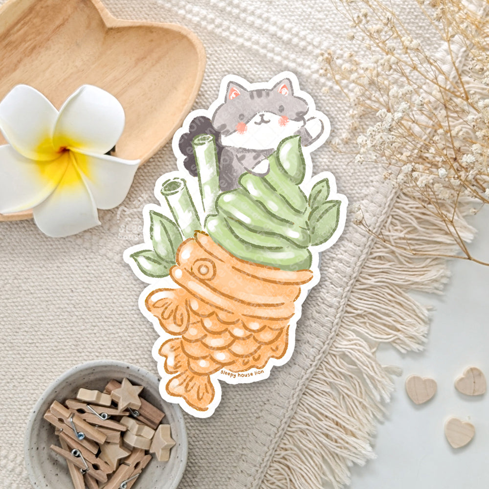 Sticker of a cat inside a Matcha Taiyaki on a decorative surface with flowers and wooden items.