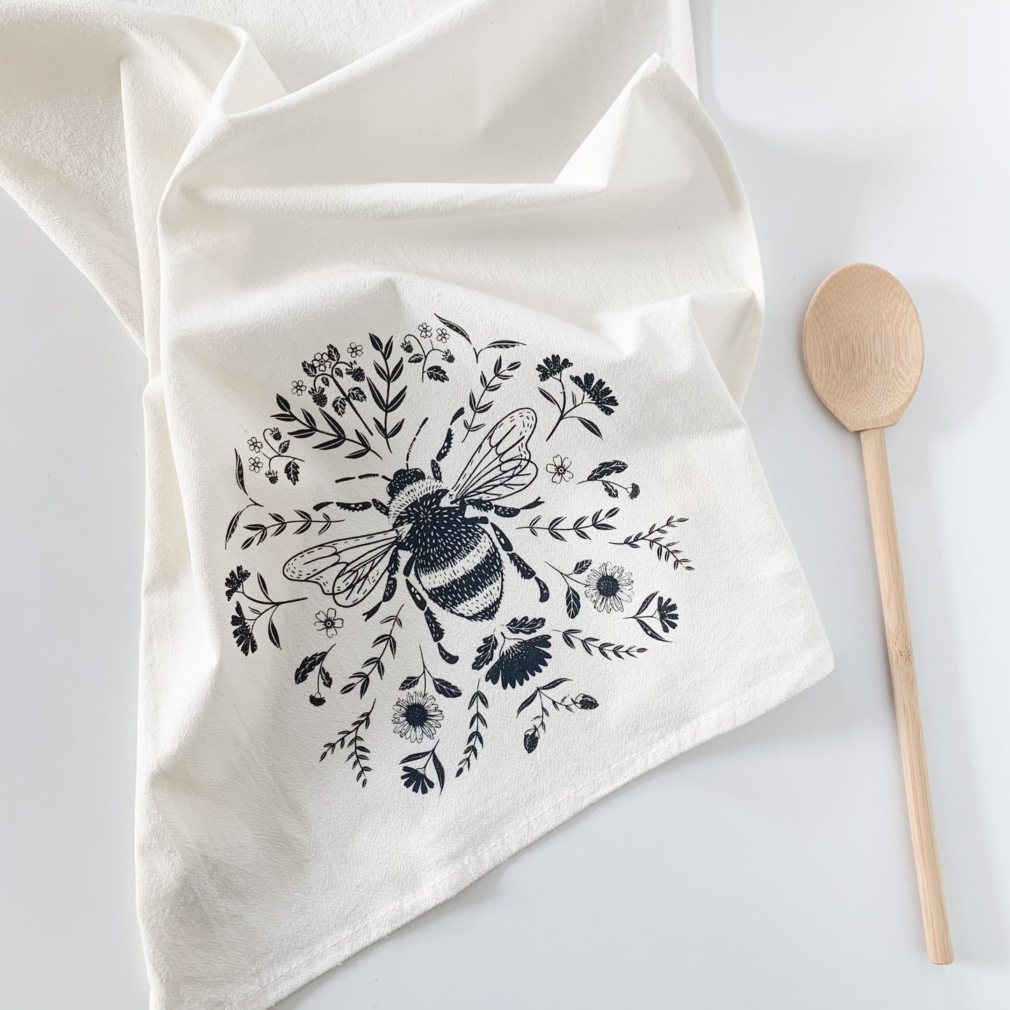 A cotton tea towel with a bee and floral pattern printed on it. There's a mixing spoon off to the side.  - Kinsfolk Shop