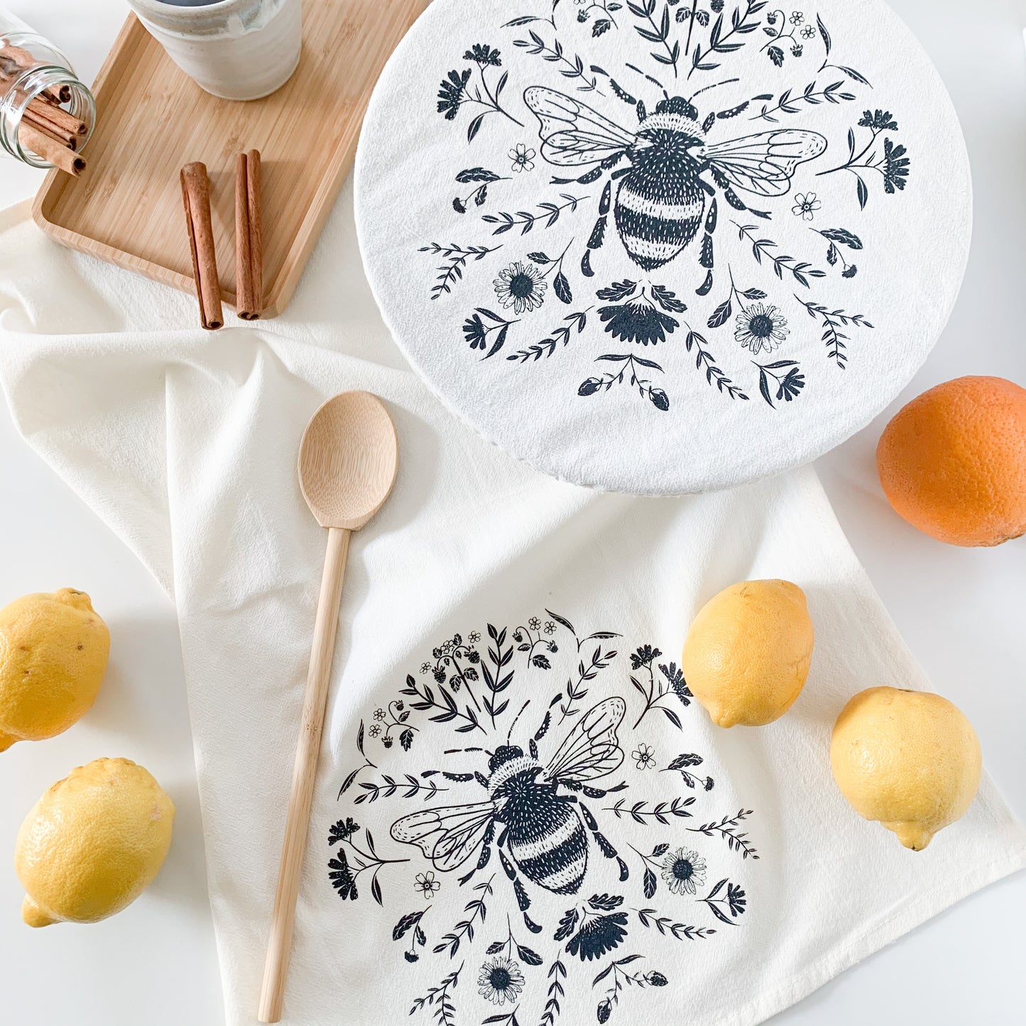A bee tea towel and matching bee cowl cover made from cotton. They're surrounded by lemons, oranges, cinnamon sticks, and bamboo utensils.
