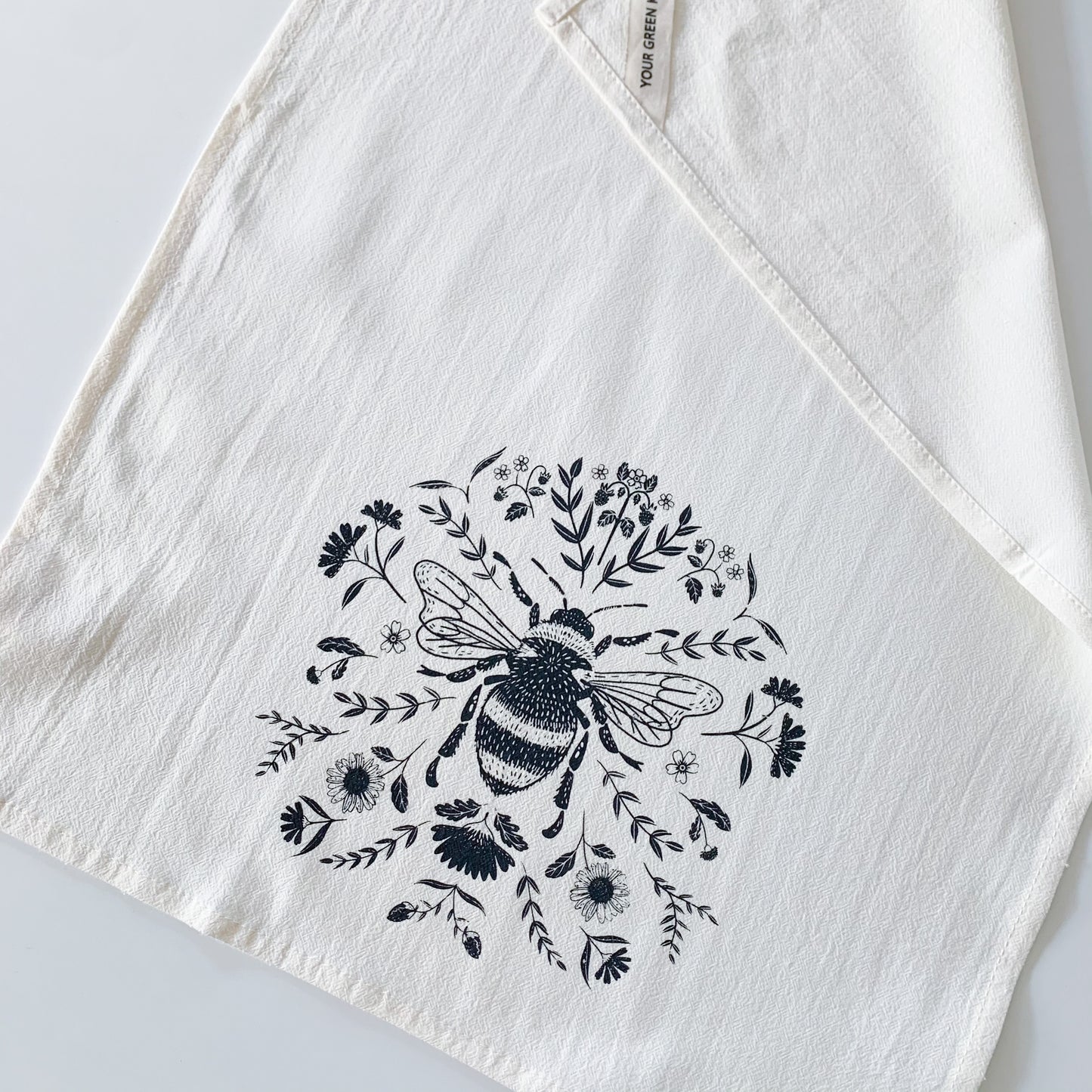 Close up of the bee and floral detail on a cotton tea towel. One of the corners of the towel if flipped over to show the hanging loop which says 'your green kitchen' on it.  - Kinsfolk Shop