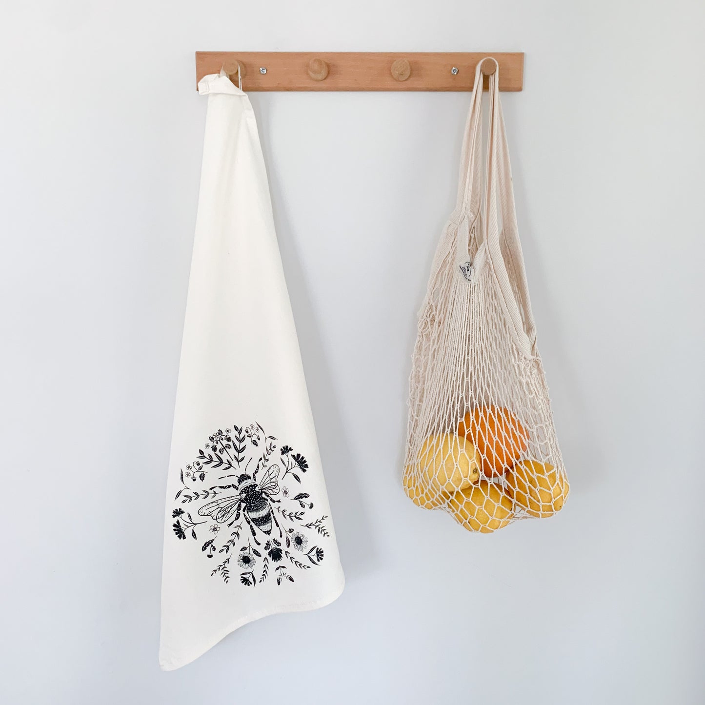 Bee Tea Towel hanging on a wood coat rack beside a cotton net tote bag which is filled with lemons and oranges.  - Kinsfolk Shop