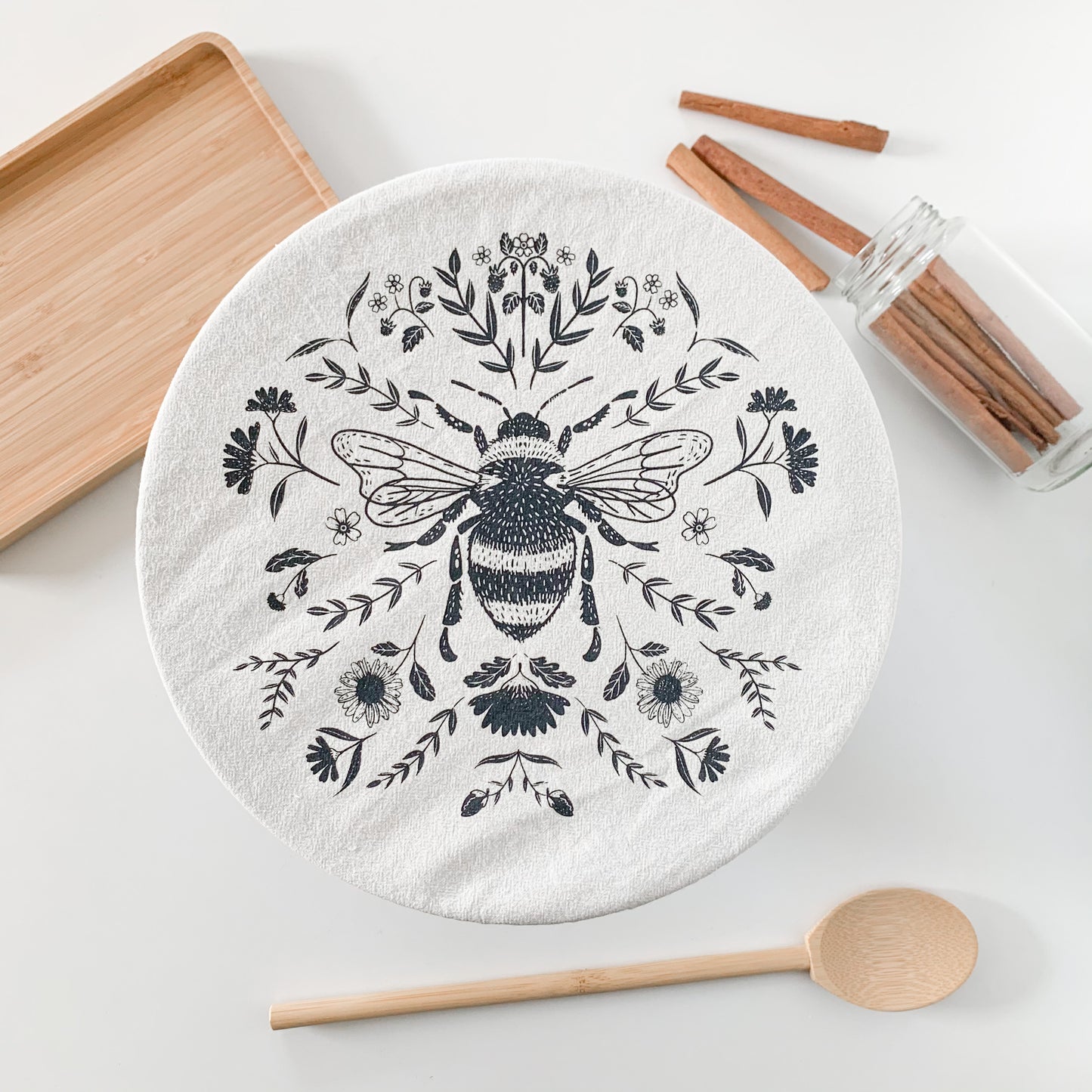 A cotton Bowl Cover with a bee and flower design printed on it. There's a mixing spoon, a small bamboo tray, and some cinnamon sticks laying around the bowl cover.